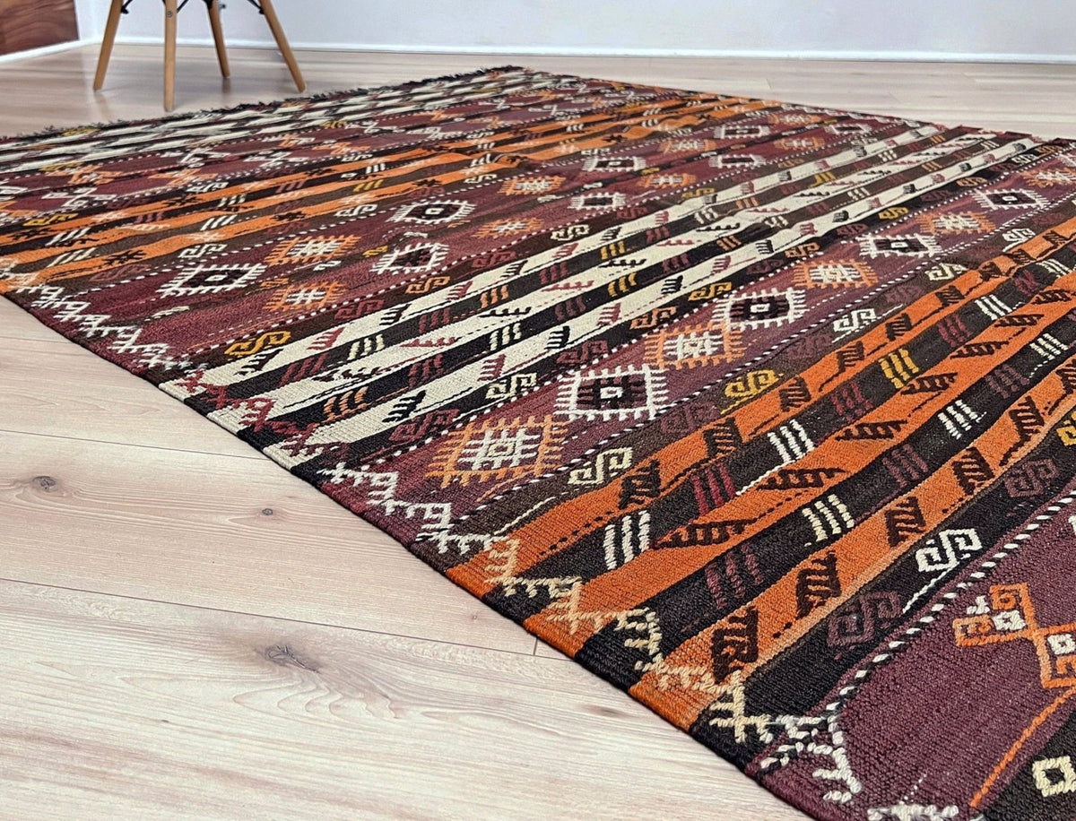 A kilim area rug with a geometric tribal pattern in rust orange, brown, burgundy, and cream, laid out on a light wood floor.
