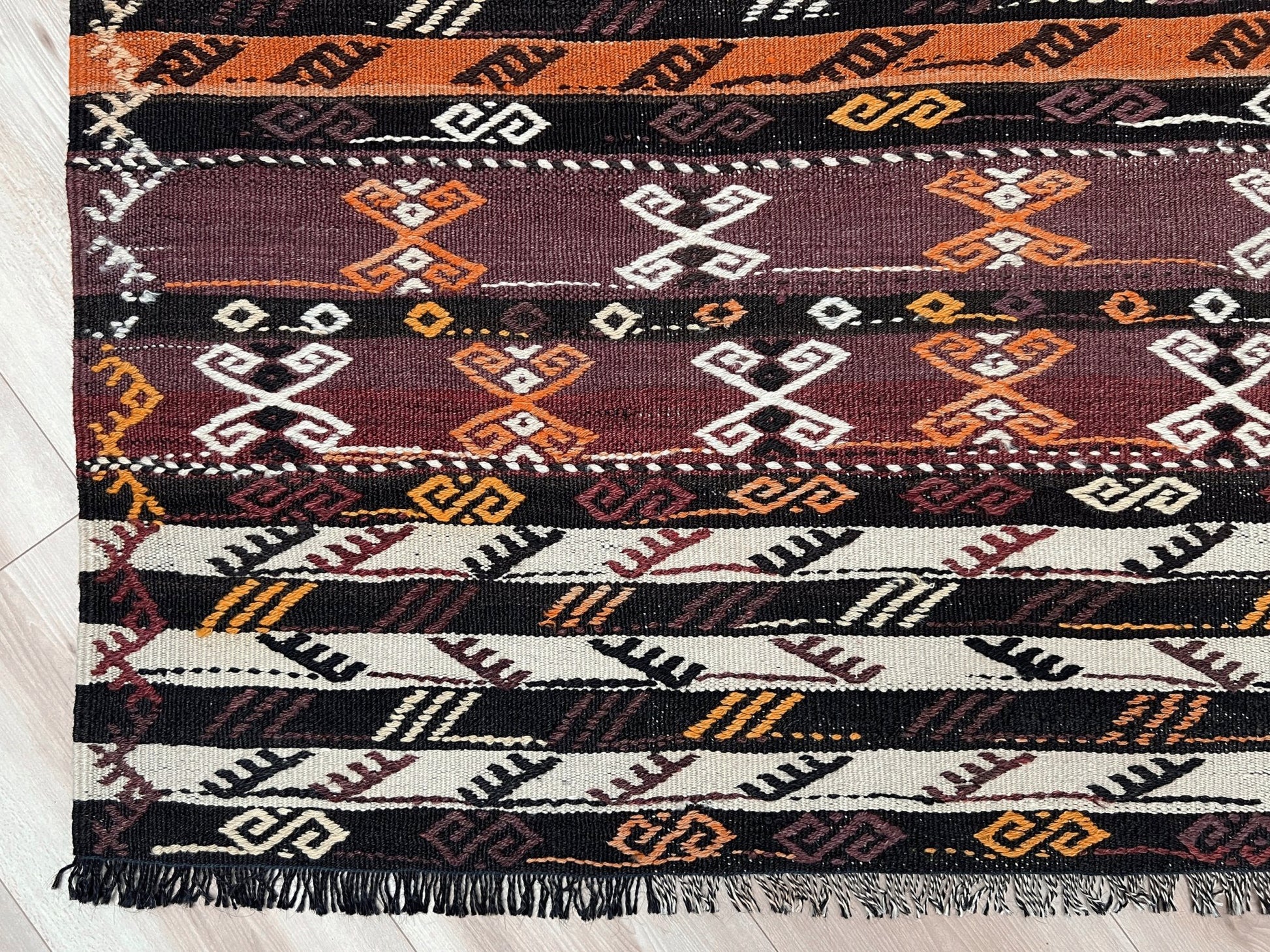 Patterned rug with geometric design on a wooden floor. Vintage Kars kilim rug from Eastern Turkey, 6x8, with horizontal tribal bands in brown, ivory, rust, and charcoal geometric motifs.