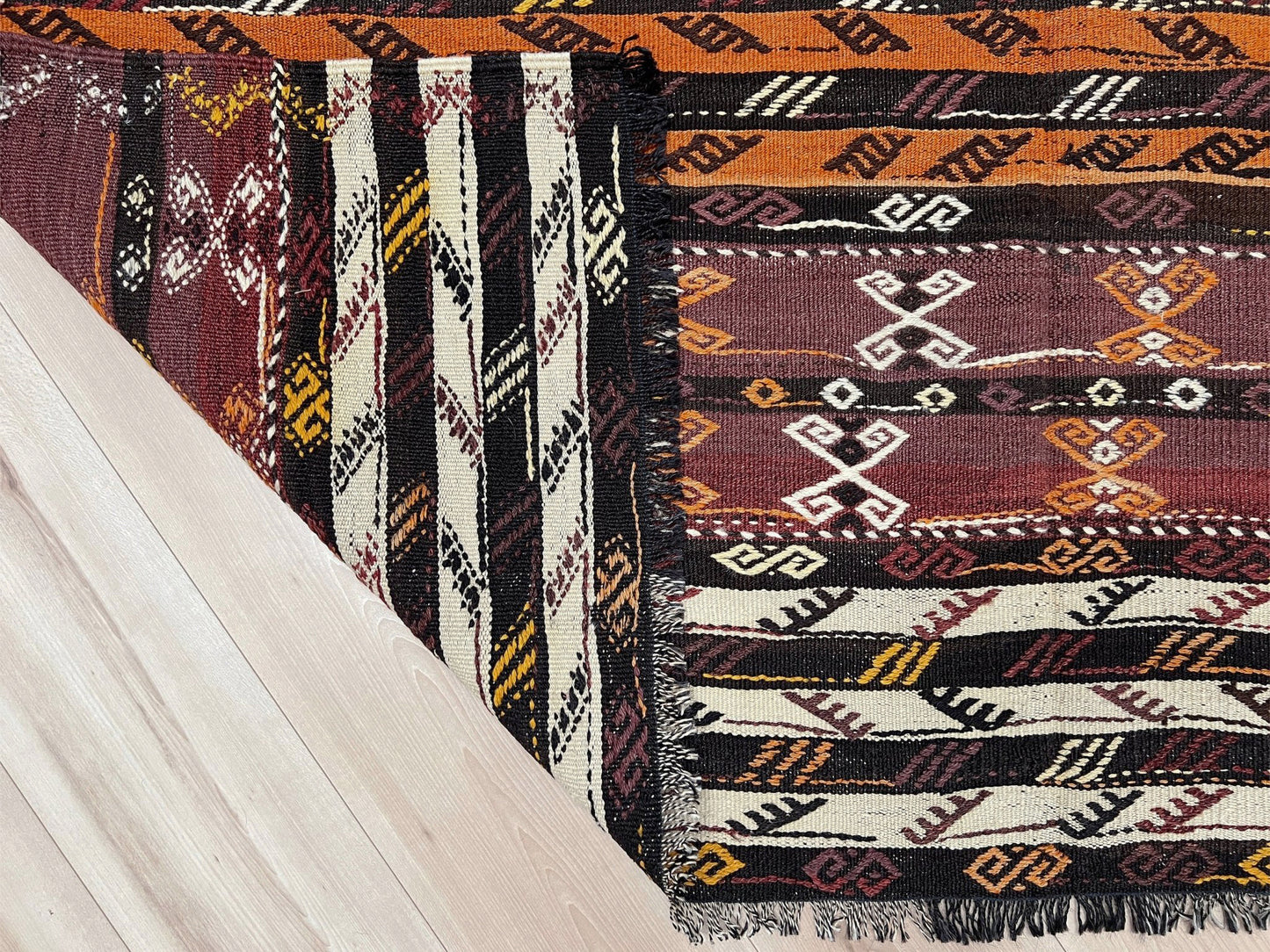 Patterned rug with geometric design on a wooden floor. Vintage Kars kilim rug from Eastern Turkey, 6x8, with horizontal tribal bands in brown, ivory, rust, and charcoal geometric motifs.
