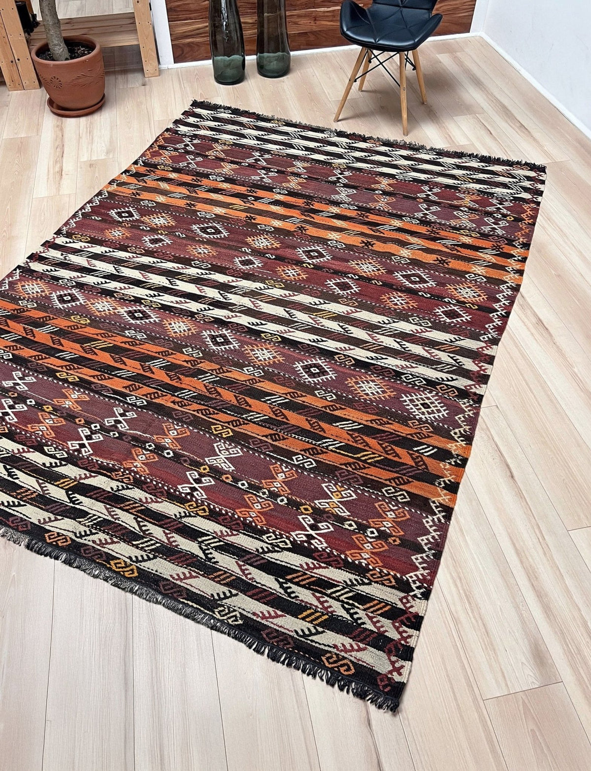 A rectangular kilim-style area rug with a geometric tribal pattern in rust orange, burgundy red, black, and cream, laid out on a light wood floor in a modern room setting.
