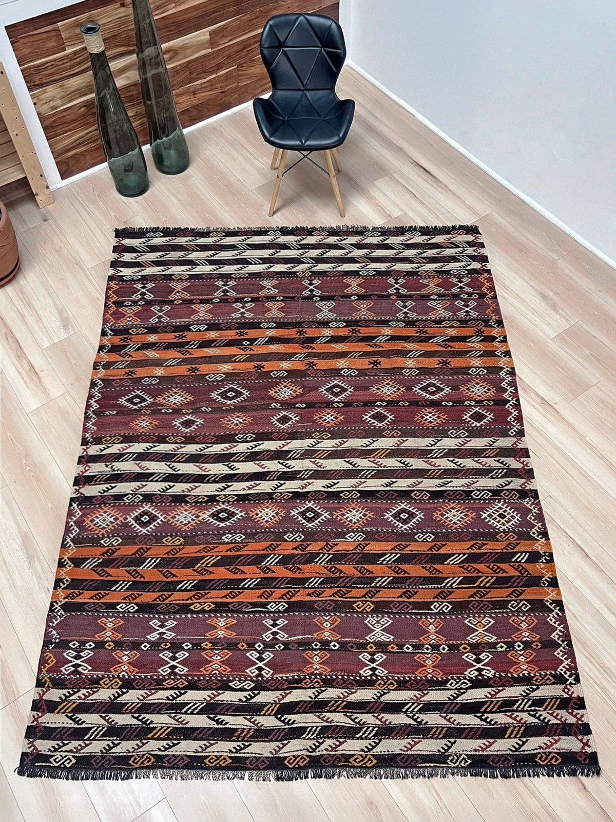 Patterned rug on a wooden floor with a chair and vases in the background. Vintage Kars kilim rug from Eastern Turkey, 6 x 8, with horizontal tribal bands in brown, ivory, rust, and charcoal geometric motifs.