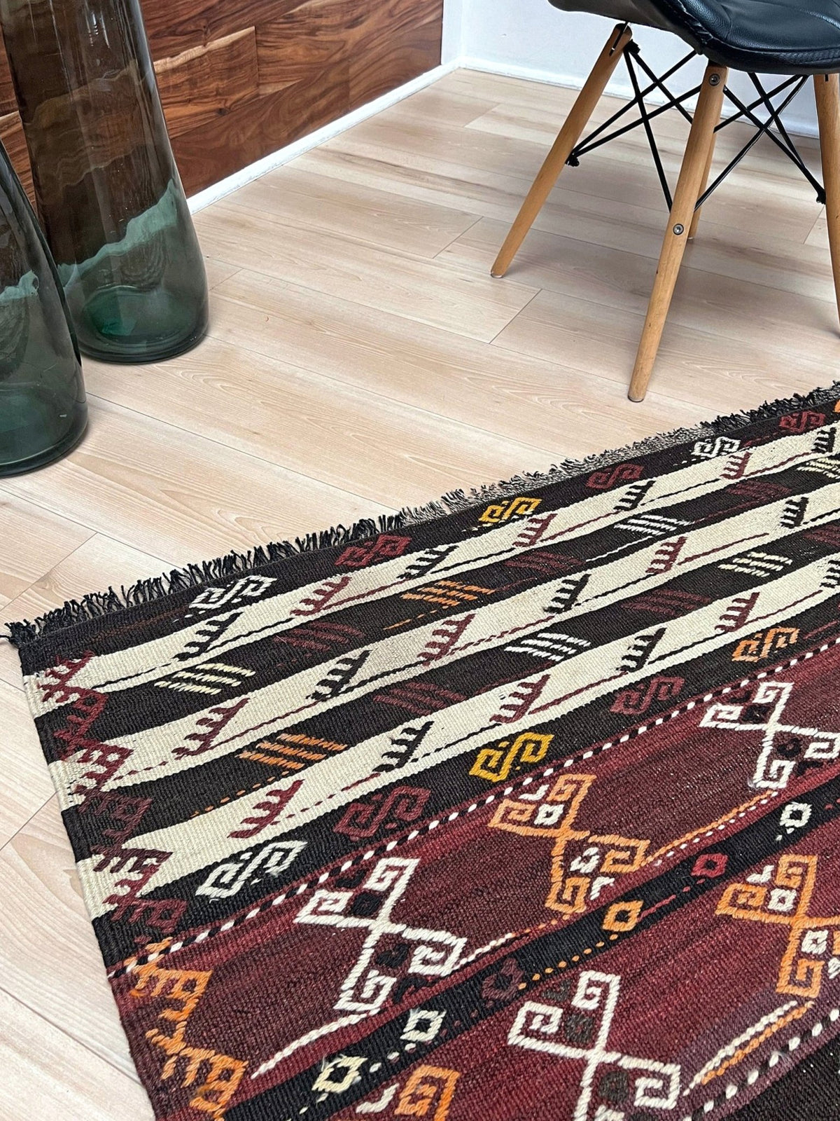 A kilim area rug with a geometric tribal pattern in red, black, beige, and orange, styled on a light wood floor in a modern room setting.