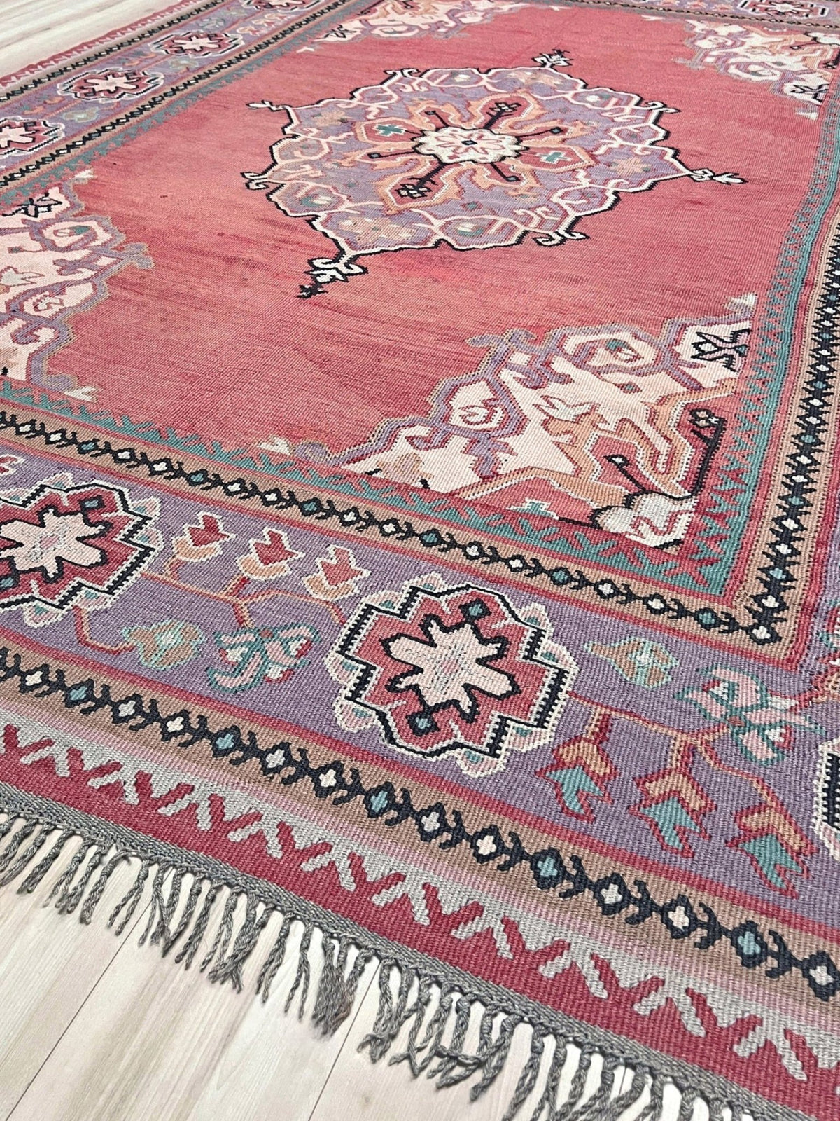 A traditional Persian-style area rug with a dusty rose pink field, featuring a central medallion, intricate geometric borders, and gray fringe tassels, laid out on a light-colored wooden floor.