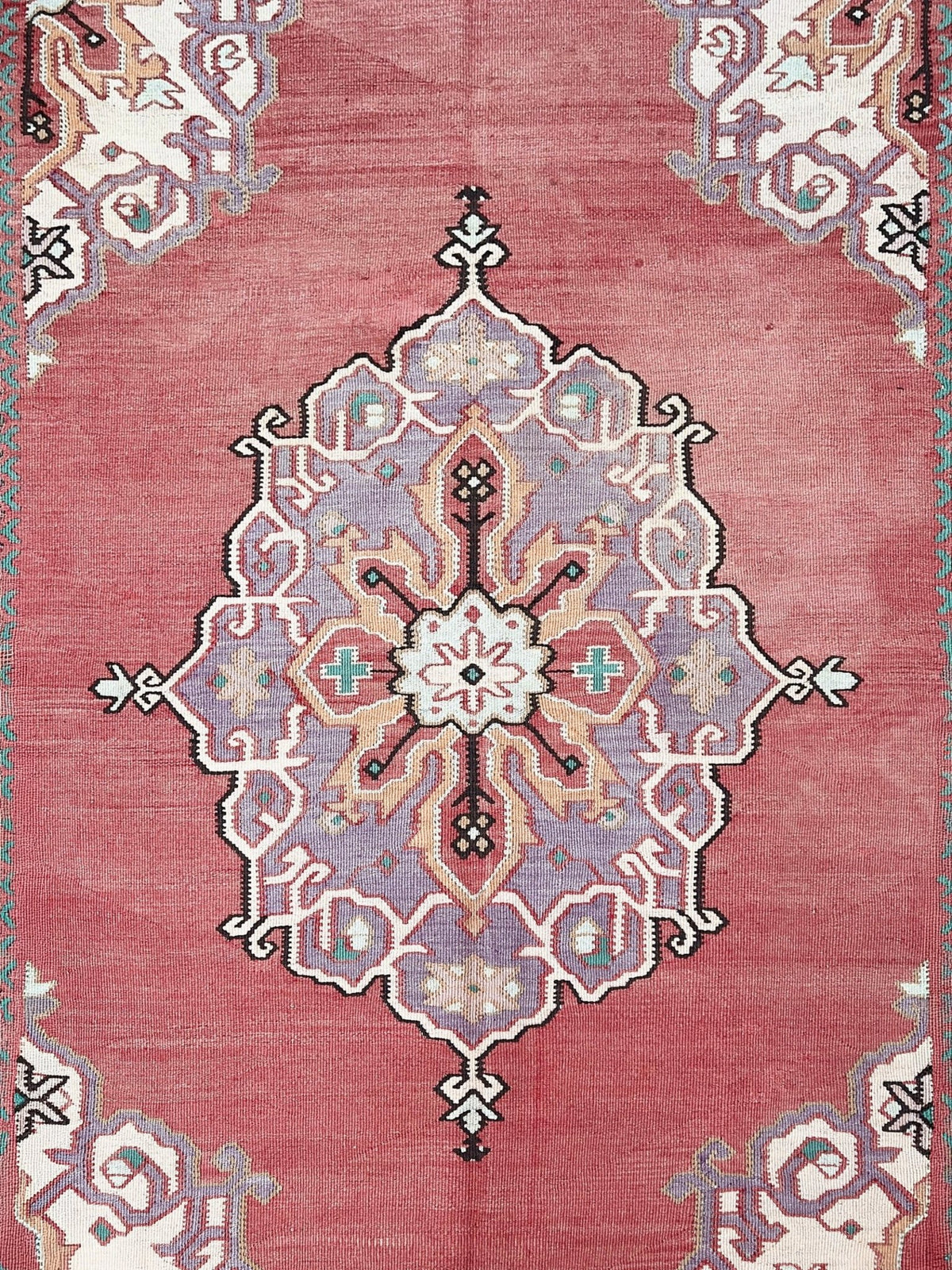 A close-up of a traditional patterned rug featuring a dusty rose pink field with a large central medallion in muted lavender, cream, black, and small turquoise accents.