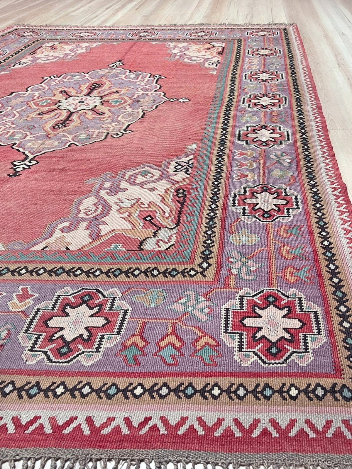 A vintage-style kilim area rug with a dusty pink field, purple and red borders, and cream floral motifs, laid out on a light wood floor.