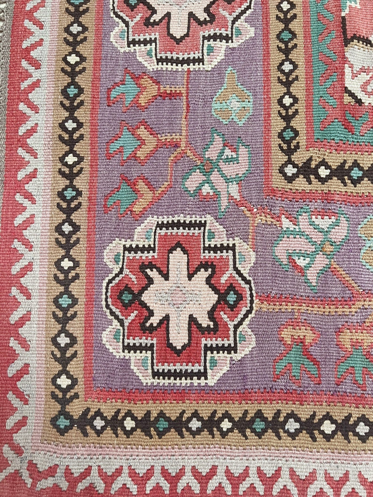 Close-up of a woven rug showing a geometric pattern with a lavender-purple field, red and cream central medallion, and multicolored floral motifs.