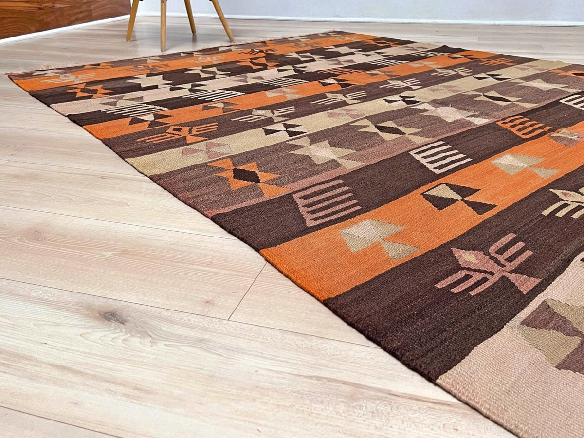 A rectangular area rug with a Southwestern geometric pattern in orange, brown, beige, and black, shown laid out on a light wood floor.