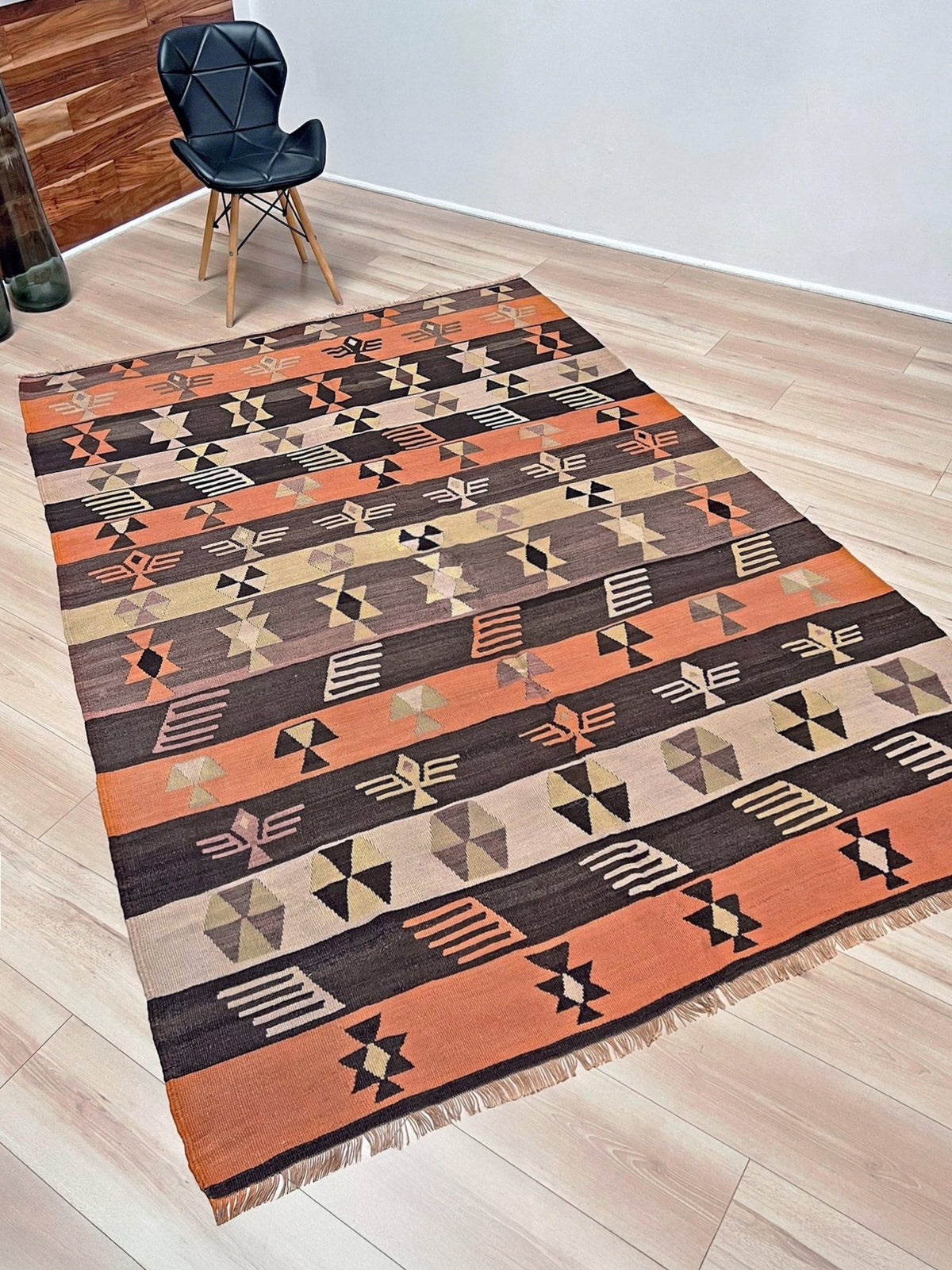 A rectangular area rug with a Southwestern geometric pattern in terracotta orange, dark brown, beige, and black, laid out on a light wood floor next to a black chair.