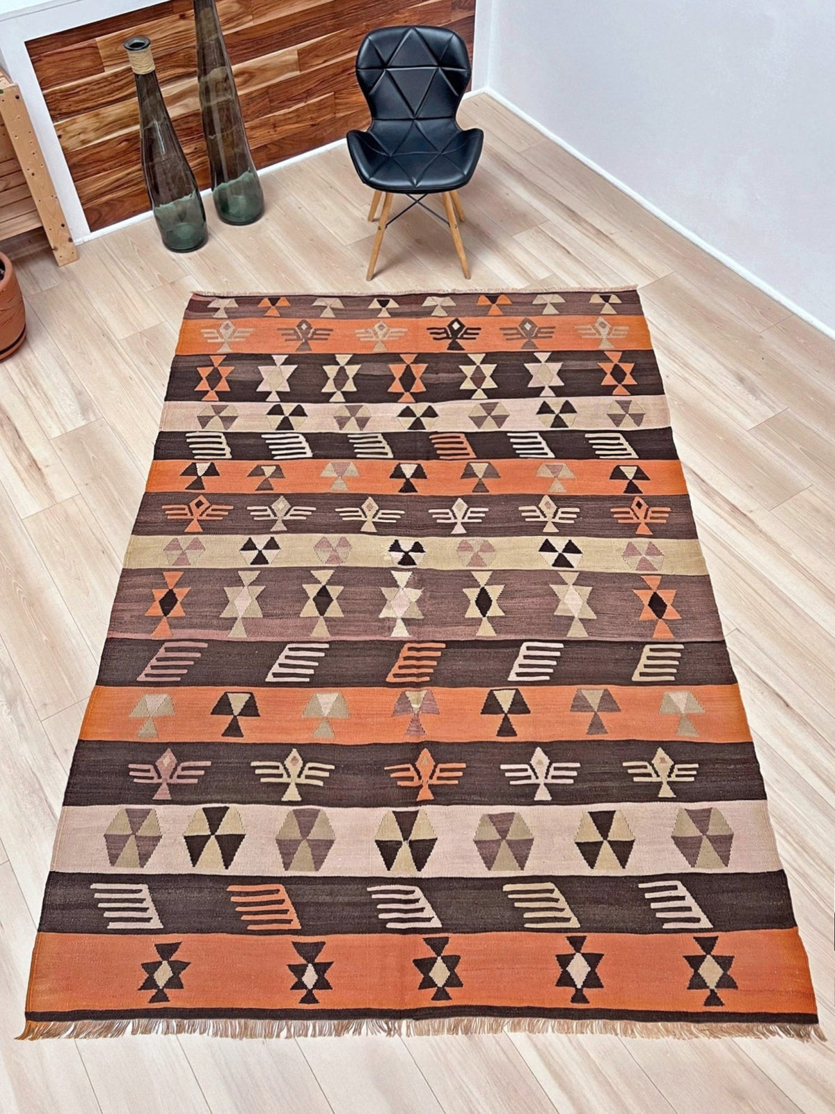 A rectangular flatweave area rug with a Southwestern geometric pattern in terracotta orange, dark brown, beige, and black, styled on a light wood floor next to a black accent chair.