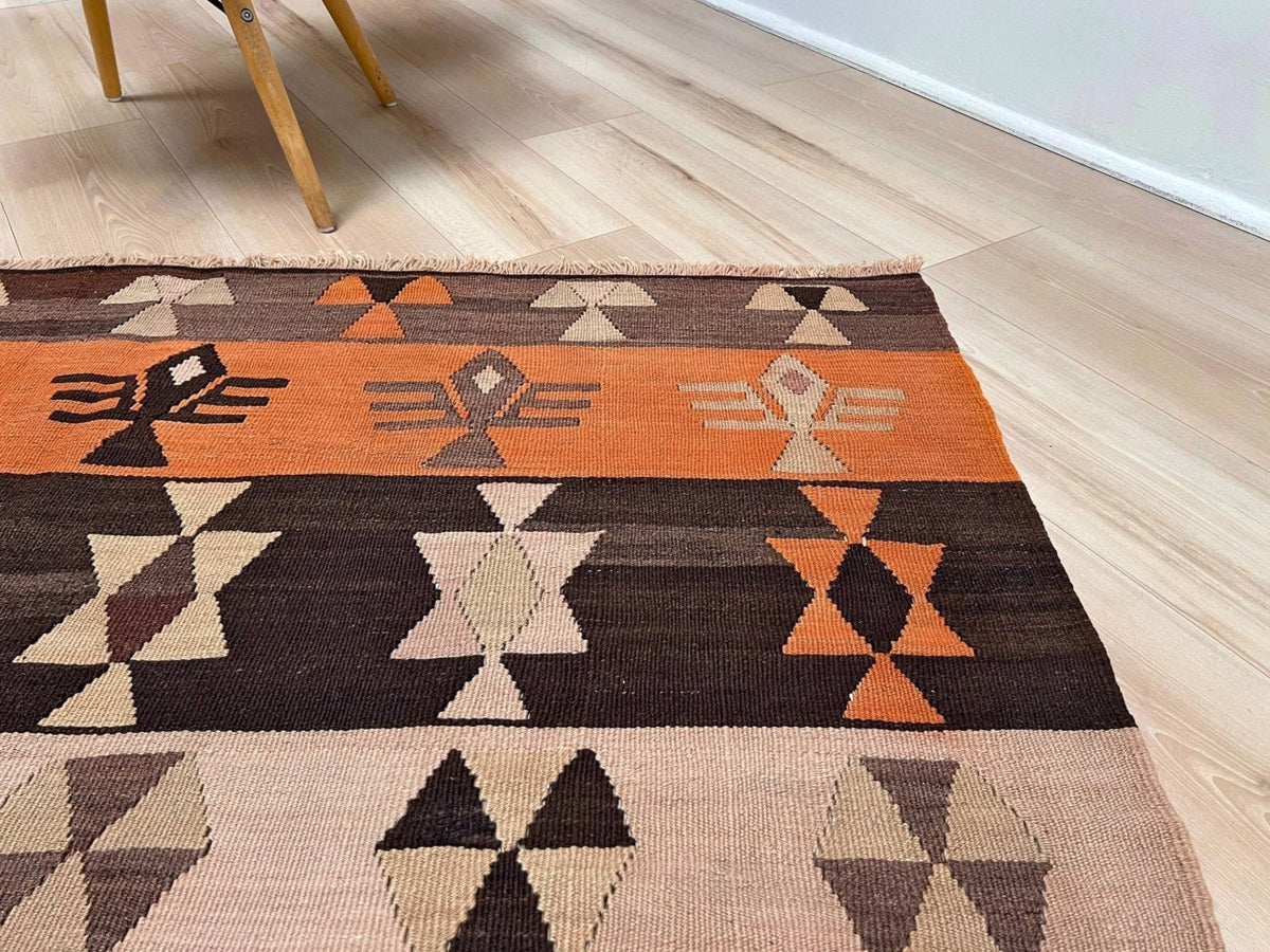 A Southwestern-style woven area rug with geometric patterns in orange, dark brown, beige, and gray, laid out on a light wood floor next to a white wall.