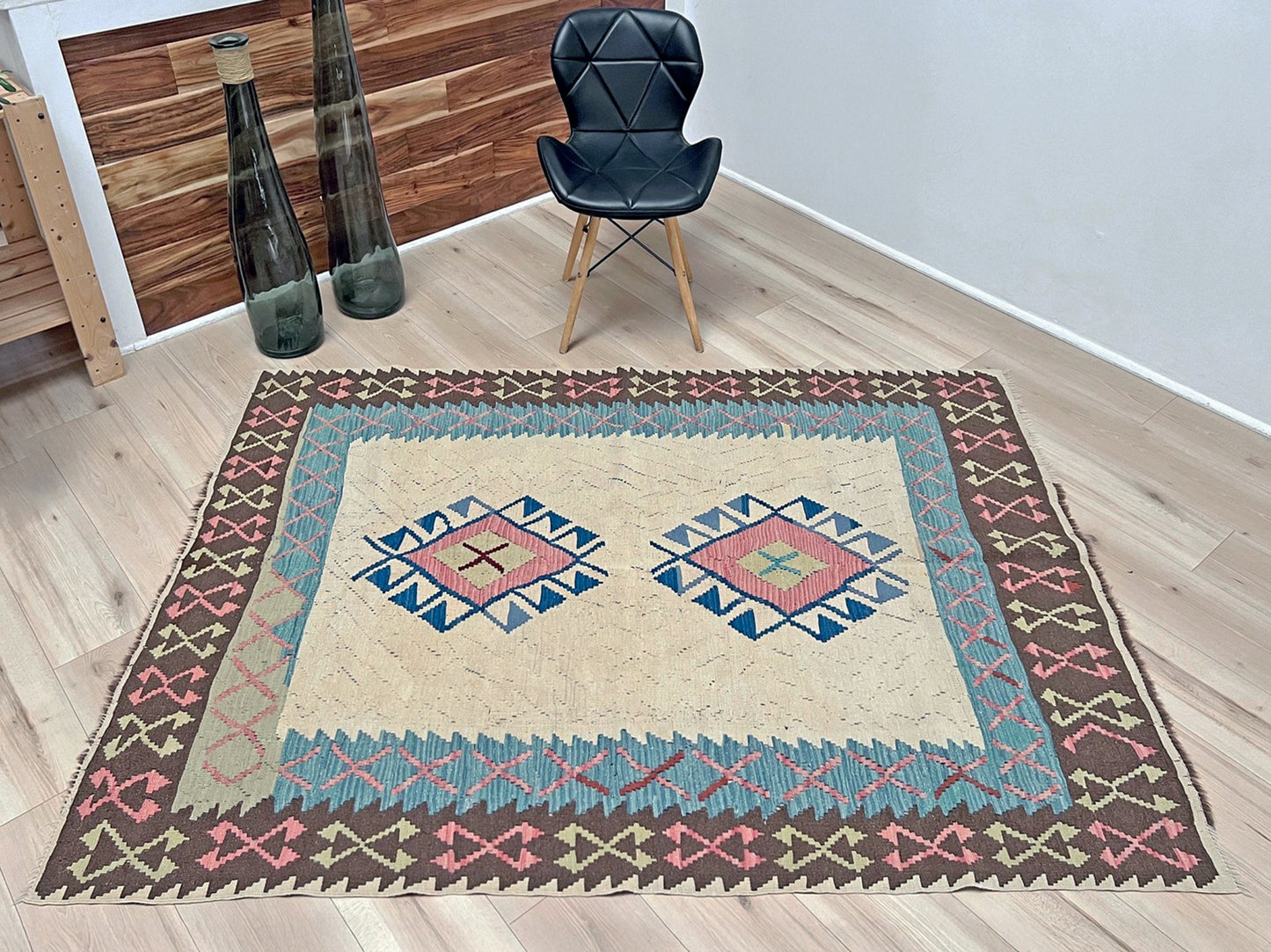 Decorative rug with geometric pattern on a wooden floor, chair, and vases in the background. Vintage Turkish kilim rug, 5x7, with ivory field, pink and blue tribal medallions, and geometric borders in brown and sage tones.