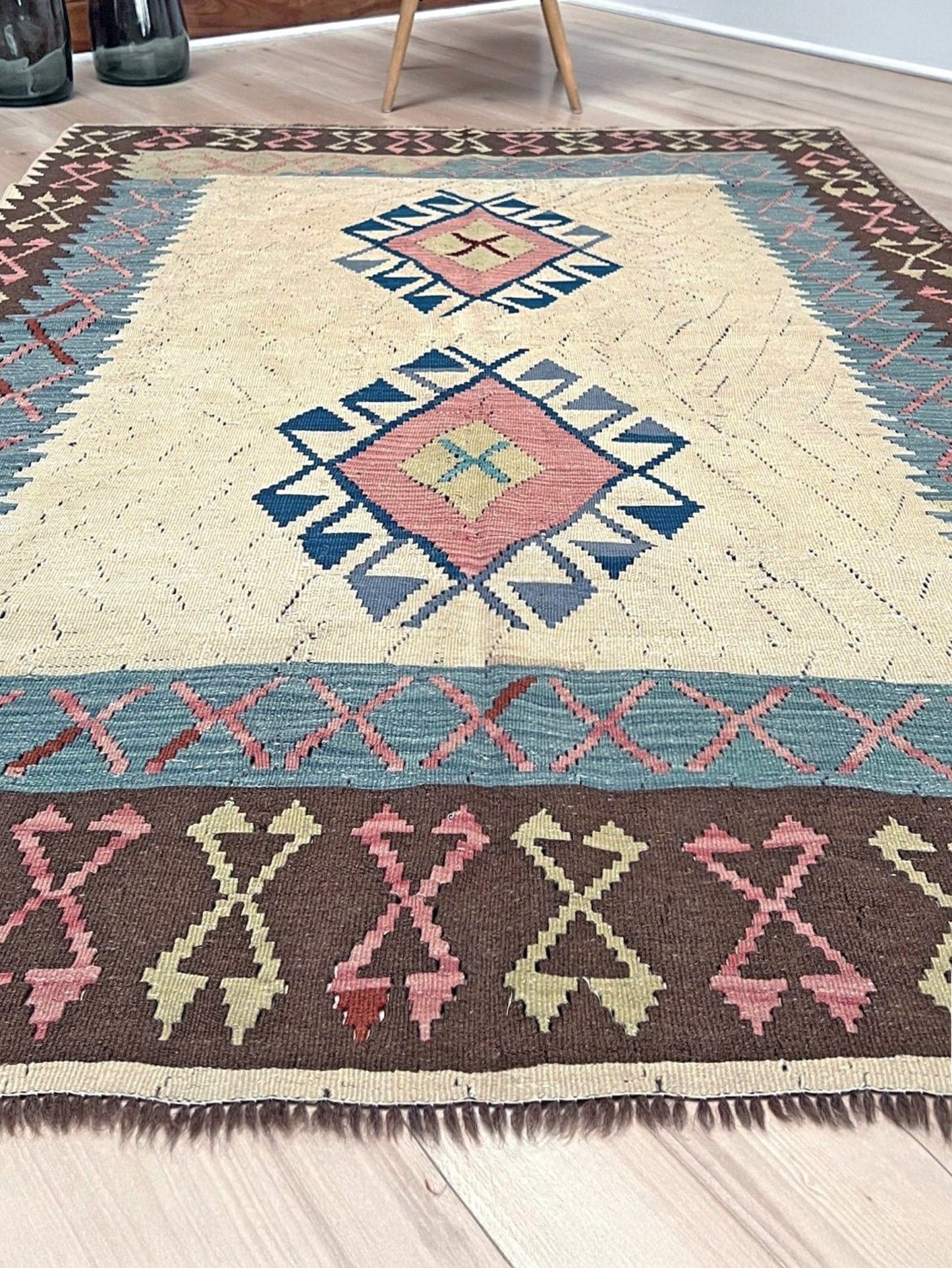 Decorative rug with geometric pattern featuring diamonds and cross symbols on a white background. Vintage Turkish kilim rug, 5x7, with ivory field, pink and blue tribal medallions, and geometric borders in brown and sage tones.