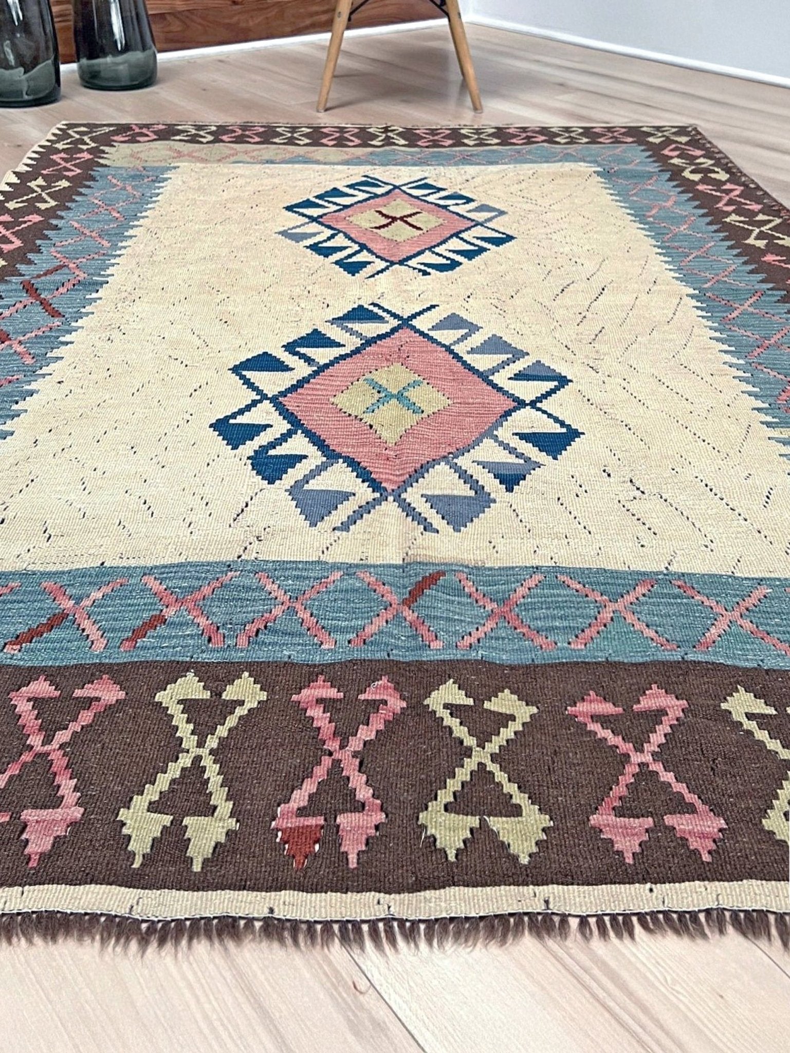 Decorative rug with geometric pattern on a wooden floor. Vintage Turkish kilim rug, 5x7, with ivory field, pink and blue tribal medallions, and geometric borders in brown and sage tones.
