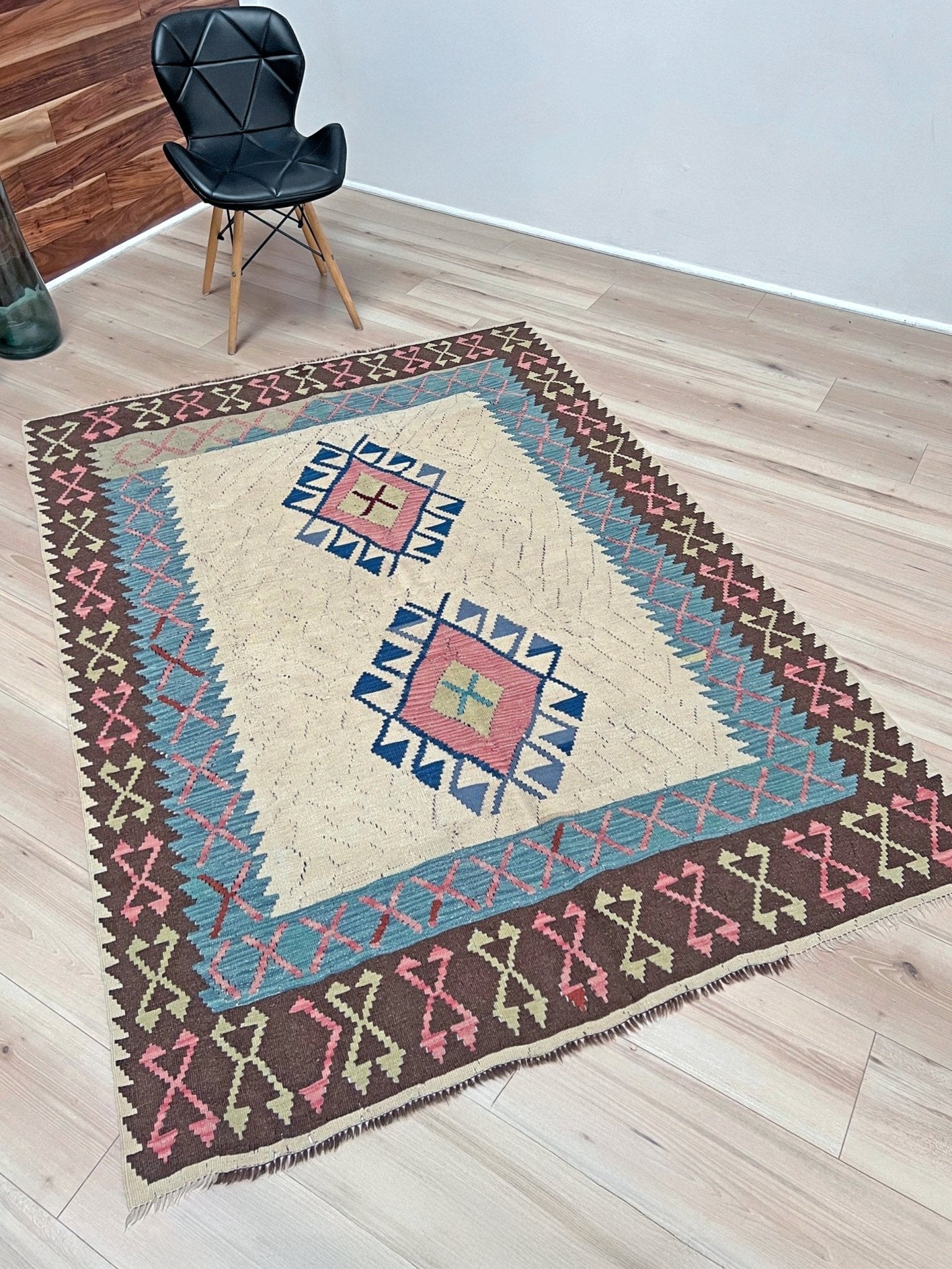 Decorative rug with geometric pattern on a wooden floor, black chair in the corner. Vintage Turkish kilim rug, 5x7, with ivory field, pink and blue tribal medallions, and geometric borders in brown and sage tones.