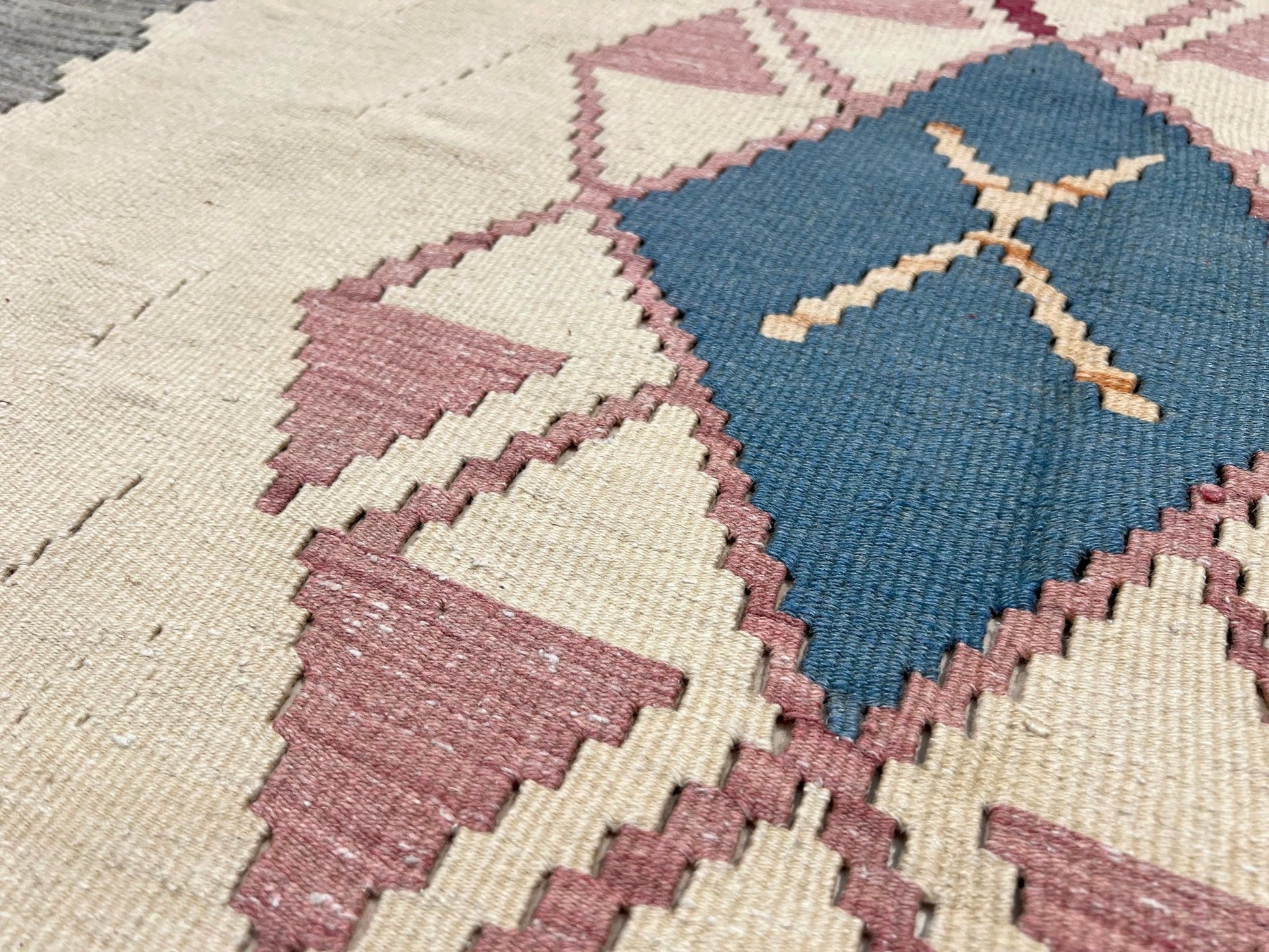 Close-up of a geometric patterned rug with blue and pink triangles on a beige background. Decorative rug with geometric pattern featuring diamonds and cross symbols on a white background. Vintage Turkish kilim rug, 5x7. 