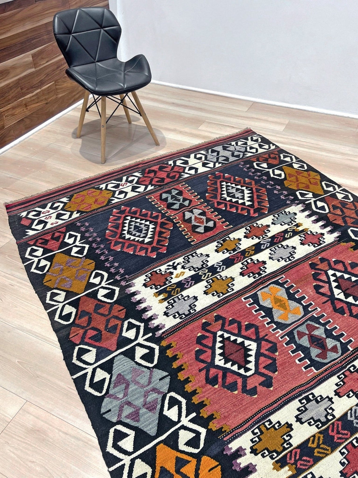 A rectangular area rug with a colorful geometric tribal pattern, shown on a light wood floor next to a black chair.