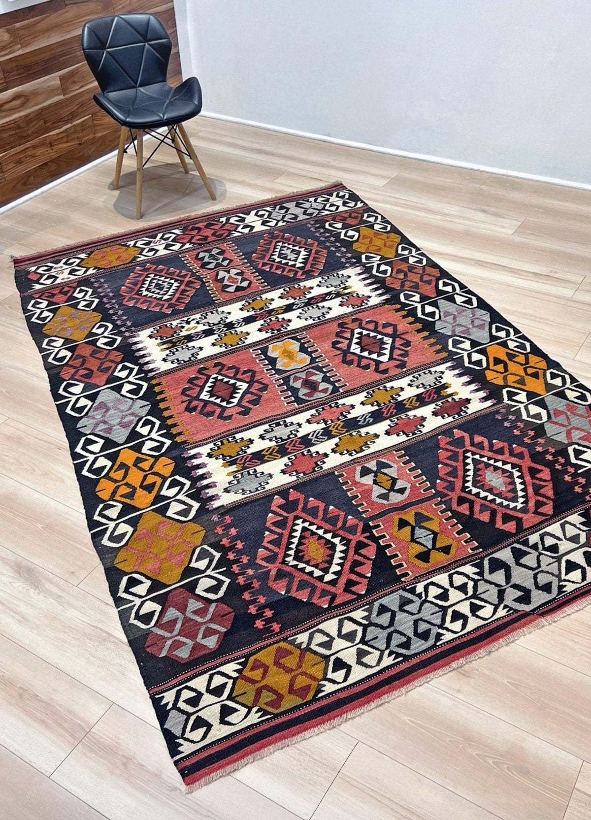 A rectangular area rug with a Southwestern geometric pattern in black, red, orange, beige, and gray, displayed on a light wood floor next to a black chair.